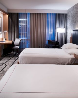 A modern hotel room with two double beds, a wall-mounted TV, a work desk with a chair, and contemporary décor.