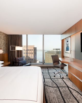 A modern hotel room with a large bed, wall-mounted TV, desk, chair, and floor-to-ceiling window overlooking buildings and water.