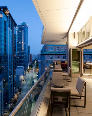 A modern cityscape at dusk, featuring tall glass buildings and a stylish balcony with tables and seating, overlooking a busy urban street.