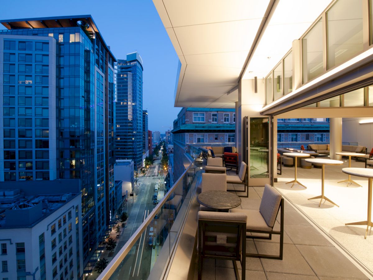 A modern cityscape view from a balcony at dusk, with outdoor seating and tables, overlooking tall buildings and a street below.