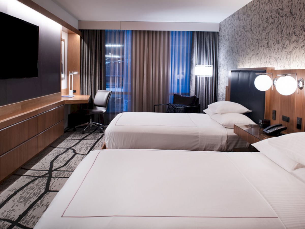 The image shows a modern hotel room with two neatly made twin beds, a desk with a chair, a flat-screen TV, and a patterned carpet.