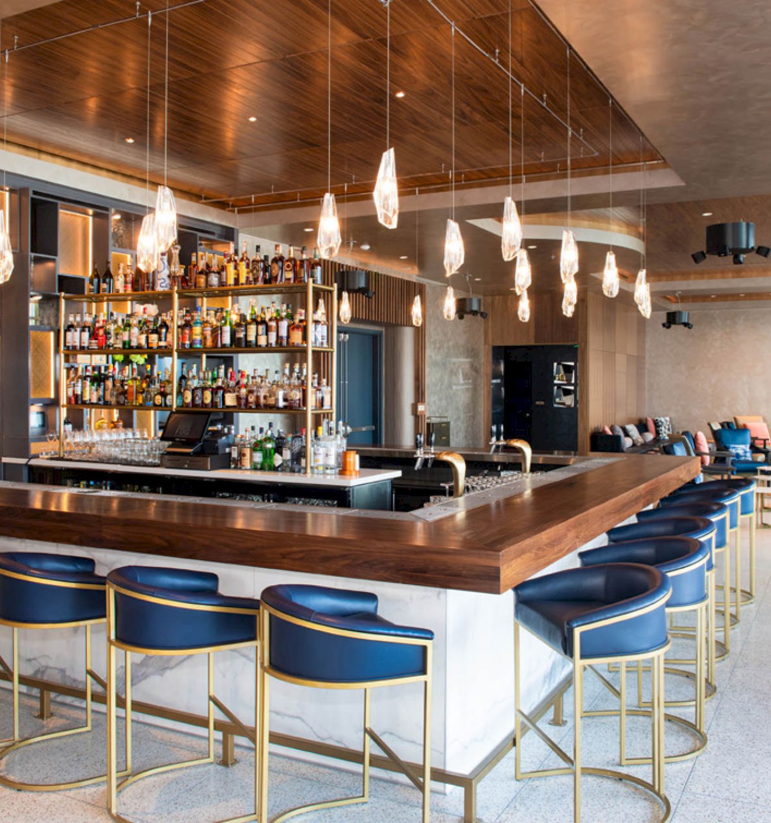 A modern bar with a long wooden counter, blue stools, a well-stocked liquor shelf, hanging lights, and a seating area in the background.