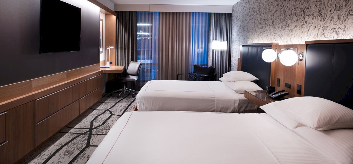 The image shows a modern hotel room with two neatly made beds, a flat-screen TV, a desk with a chair, and contemporary decor.