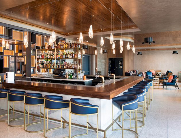 The image shows a modern bar area with stylish seating, a well-stocked bar, hanging lights, and contemporary decor, featuring lounge seating in the background.