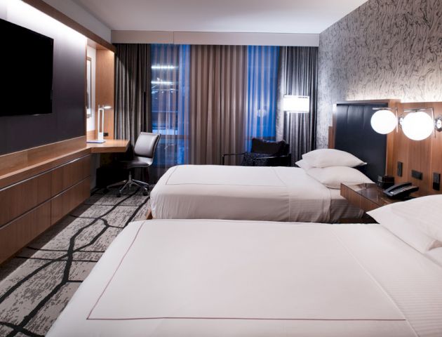 A modern hotel room with two beds, a desk area, a flat-screen TV, and stylish decor. The room features large windows with curtains ending the sentence.