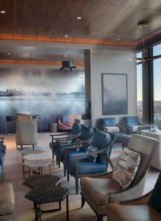 A modern lounge with various seating options and large windows offering a view of a waterfront, ferris wheel, and vibrant cityscape outside.