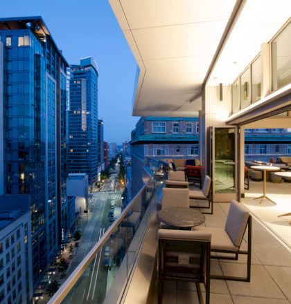 A modern cityscape at dusk, with a view from a high-rise building's balcony furnished with chairs and tables and neighboring buildings in the background.
