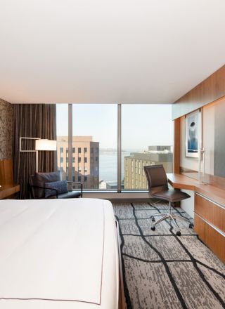 A modern hotel room with a large bed, a flat-screen TV, a desk with a chair, and a view of buildings and water through the window.
