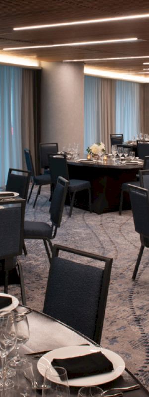 The image shows an elegantly set dining area with round tables, chairs, glassware, and centerpieces, all under soft overhead lighting.