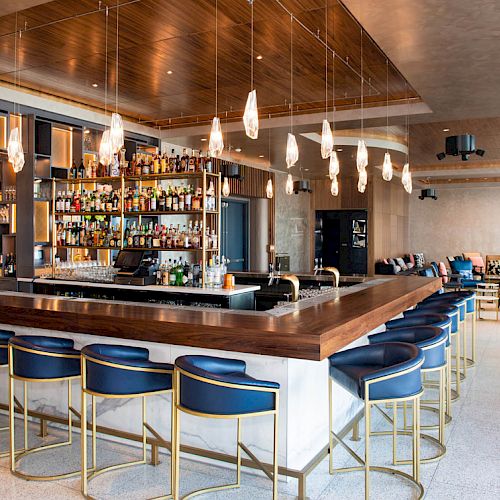 A modern bar with a wooden countertop, blue stools, hanging light fixtures, a large selection of spirits, and a seating area in the background.