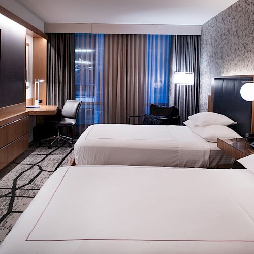 A modern hotel room features two neatly made beds, a wall-mounted TV, a desk with a chair, and contemporary lighting.