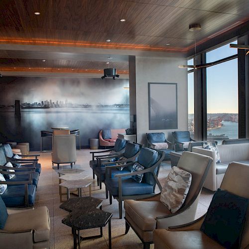 This image shows a luxurious lounge area with modern chairs, panoramic windows offering a waterfront view, and contemporary artwork on the back wall.