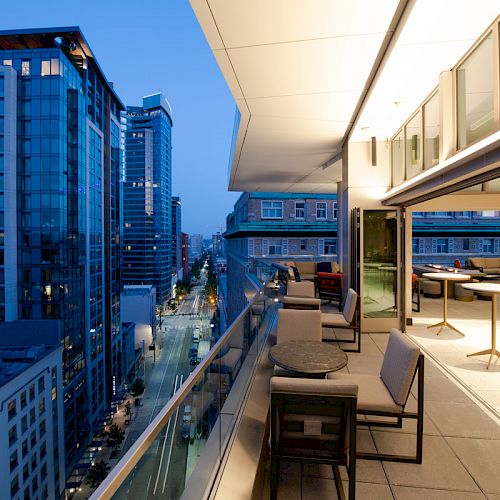 A modern balcony at dusk overlooks an urban street with tall buildings, featuring comfortable seating and tables under soft lighting.