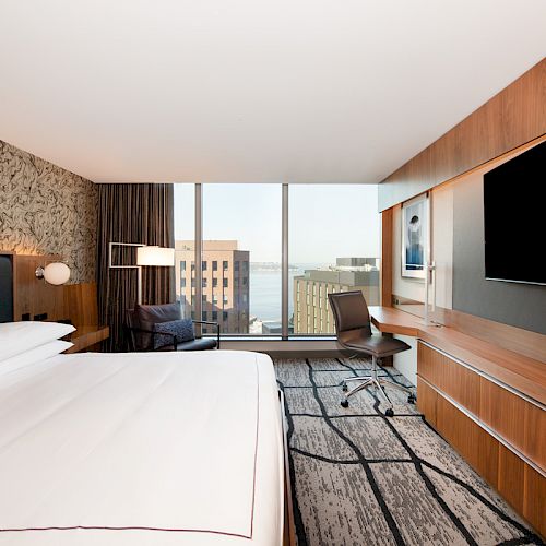 A modern hotel room with a large bed, a TV, a desk with a chair, an armchair, and a view of buildings and water through large windows.