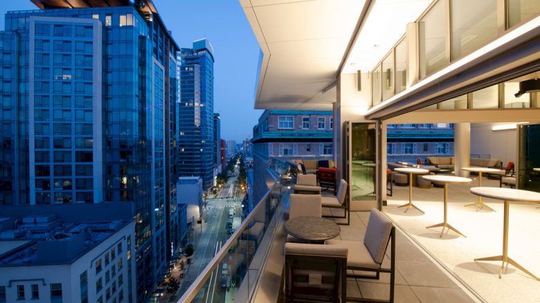 A modern balcony with several tables and chairs overlooking a cityscape of tall buildings at dusk, with streetlights illuminating the scene.
