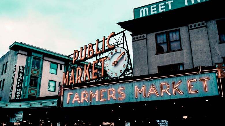 The image shows an iconic market sign with "PUBLIC MARKET" and "FARMERS MARKET," along with a clock, in an urban setting with various buildings.