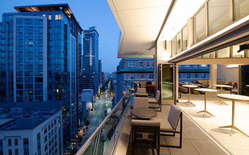 The image shows a modern balcony with tables and chairs, overlooking a city street and tall buildings at dusk, with bright interior lighting.