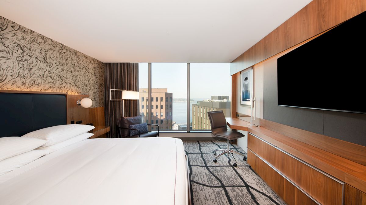 A modern hotel room with a large bed, a desk with a chair, a flatscreen TV, and a view of city buildings through the large window.