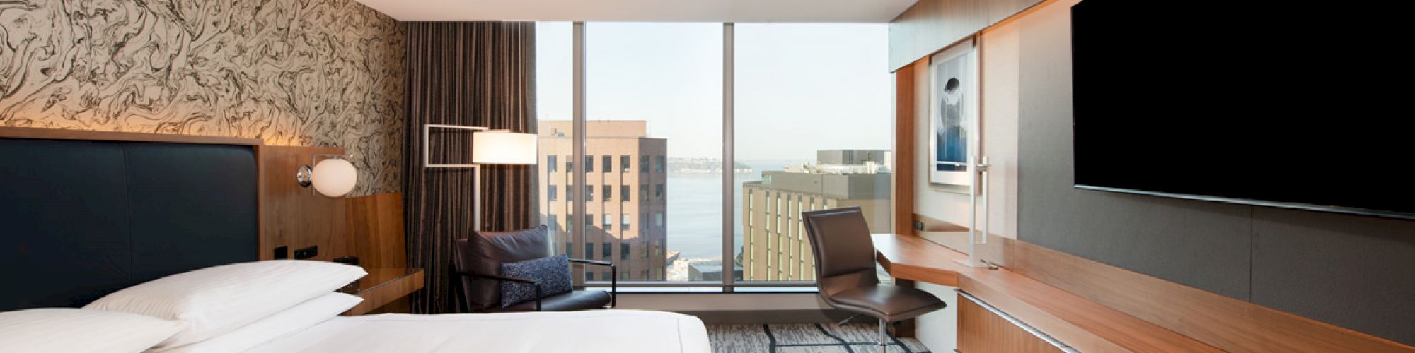 A modern hotel room with a large bed, a desk with a chair, a flatscreen TV, and a view of city buildings through the large window.