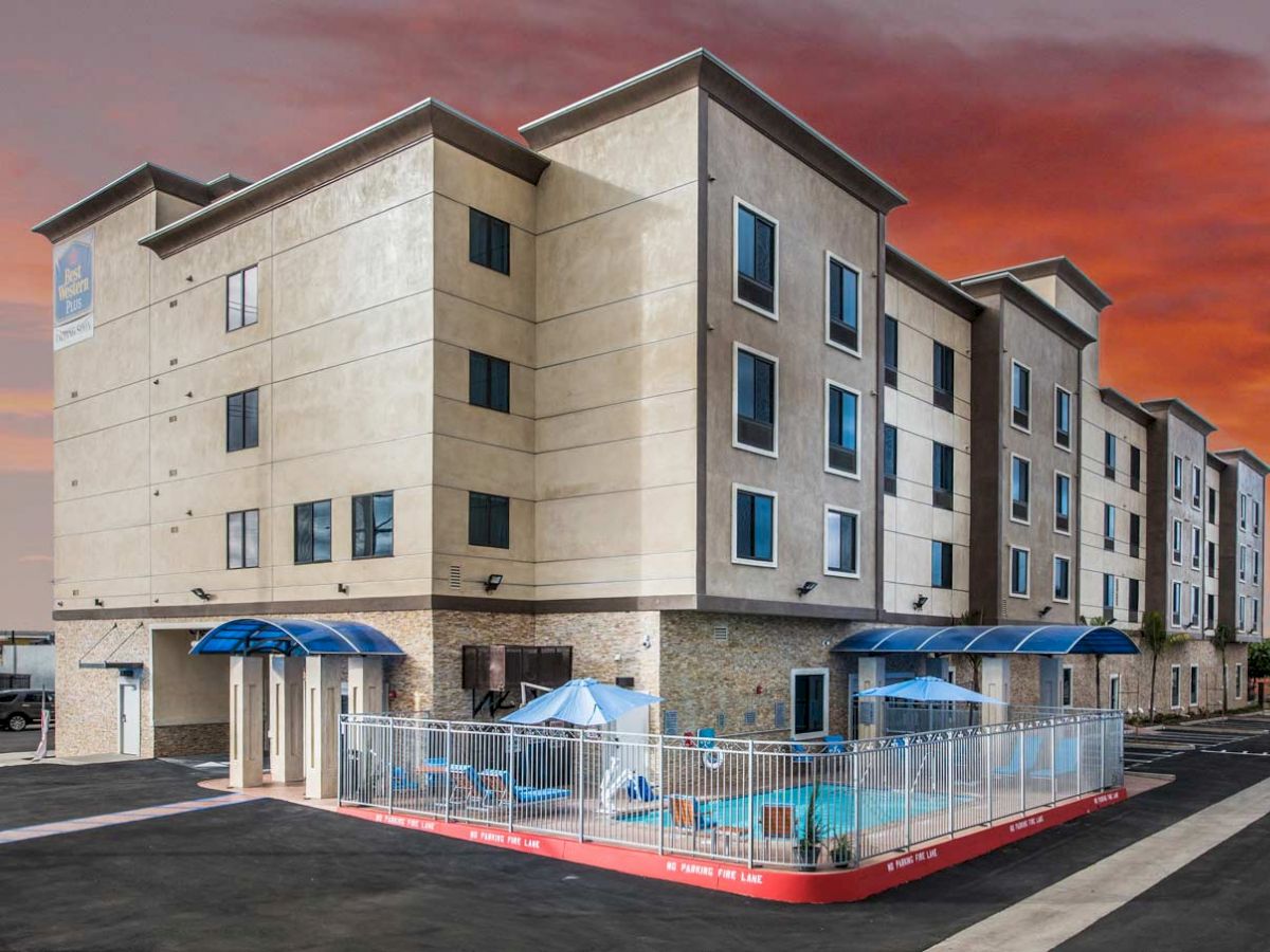 The image shows a four-story building with an outdoor pool area, surrounded by a fence. The sky is painted with a vivid sunset.