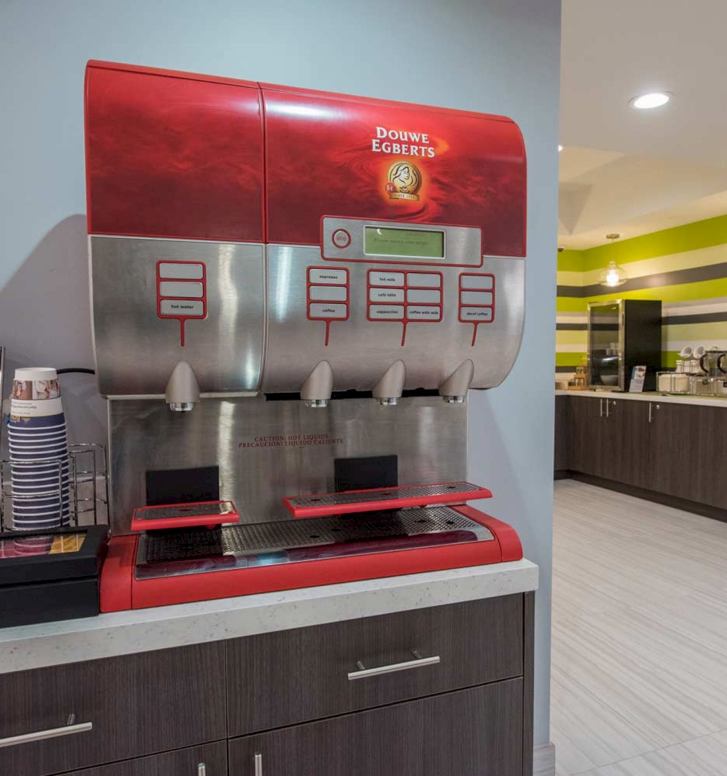 The image shows a modern beverage station with a Dunkin' Donuts coffee machine, cups, condiments, and a snack holder on a counter in a stylish room.