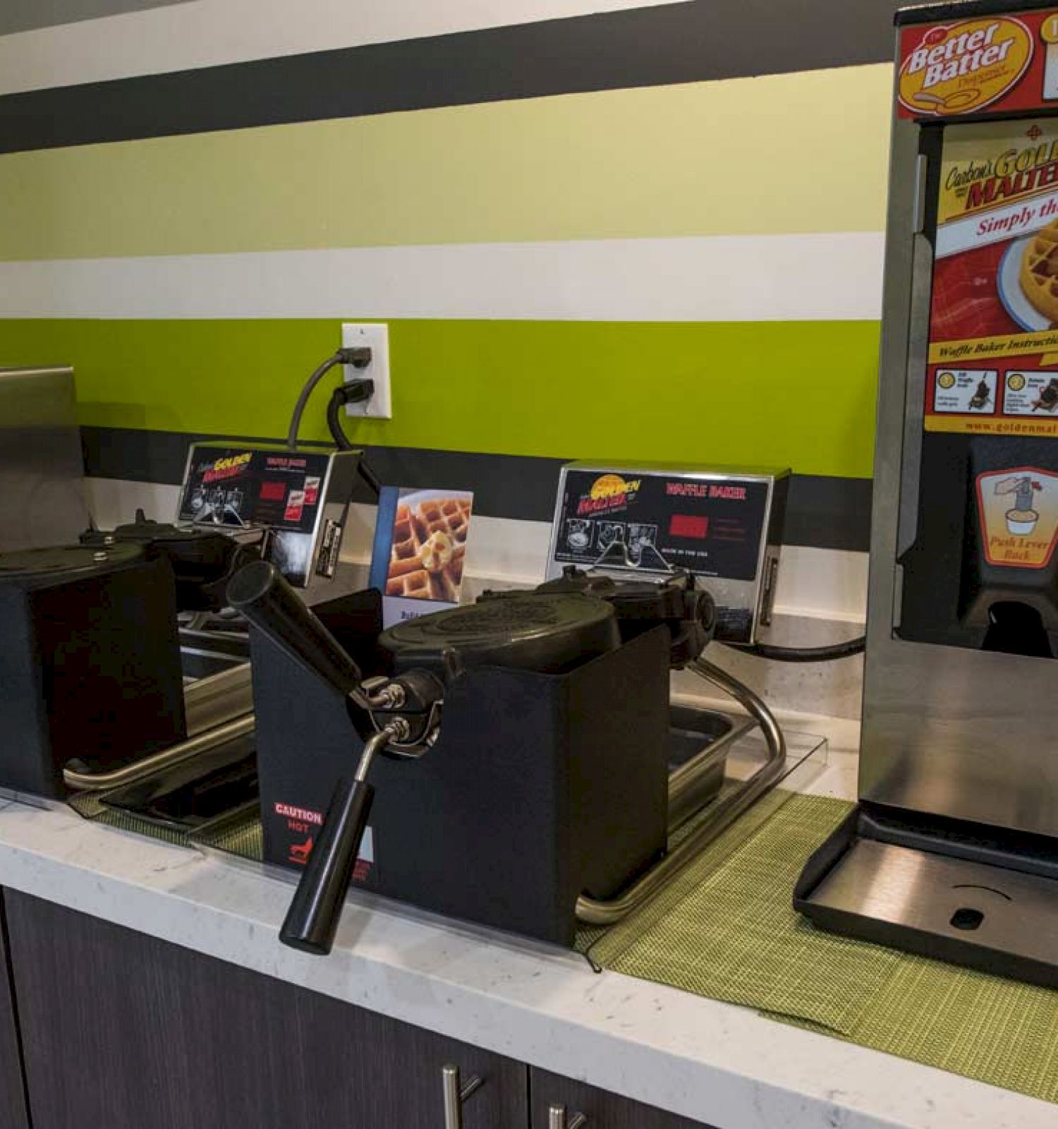 The image shows a breakfast station with waffle makers, a waffle batter dispenser, a microwave, and syrup containers on a countertop.