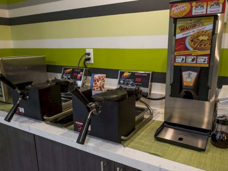 The image shows a breakfast station with waffle makers, a microwave, and a waffle mix dispenser, accompanied by syrup bottles and napkins.