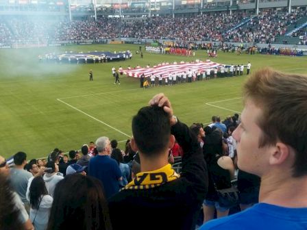 A crowd is gathered in a stadium, watching a large flag being displayed on the field, with people standing around it.