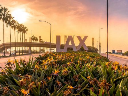 The image shows the iconic LAX sign at Los Angeles International Airport with a backdrop of palm trees and a sunset sky.