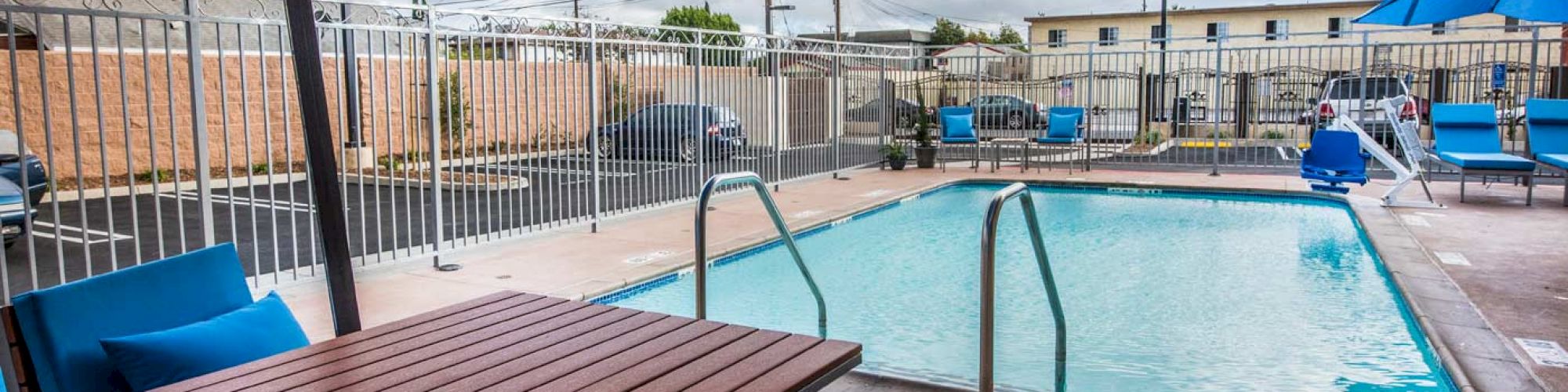 This image shows a small outdoor pool area with blue umbrellas, lounge chairs, tables, and fencing surrounding the pool. The weather is partly cloudy.