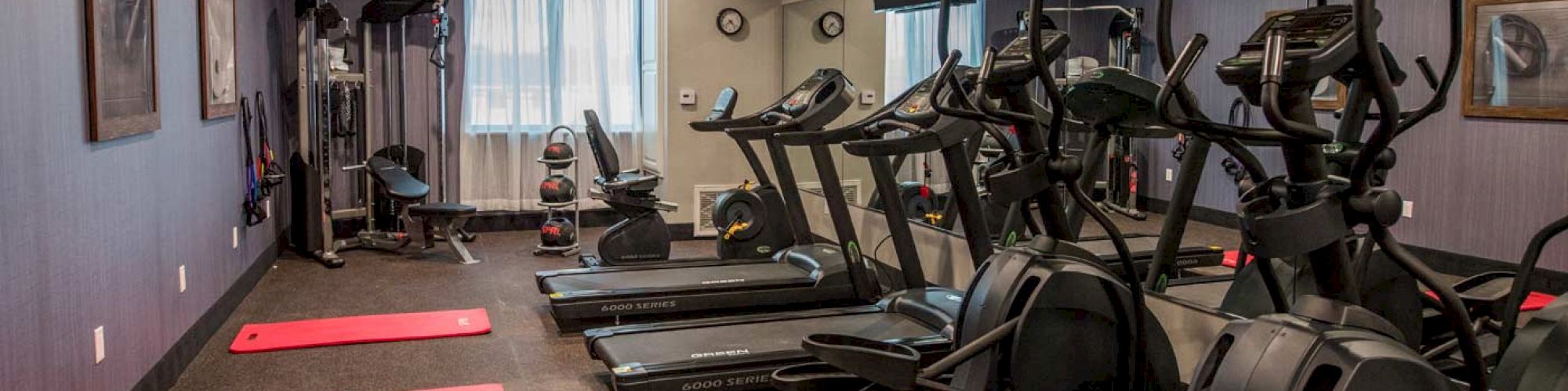 A modern gym with treadmills, elliptical machines, red exercise mats, weightlifting equipment, and wall mirrors.