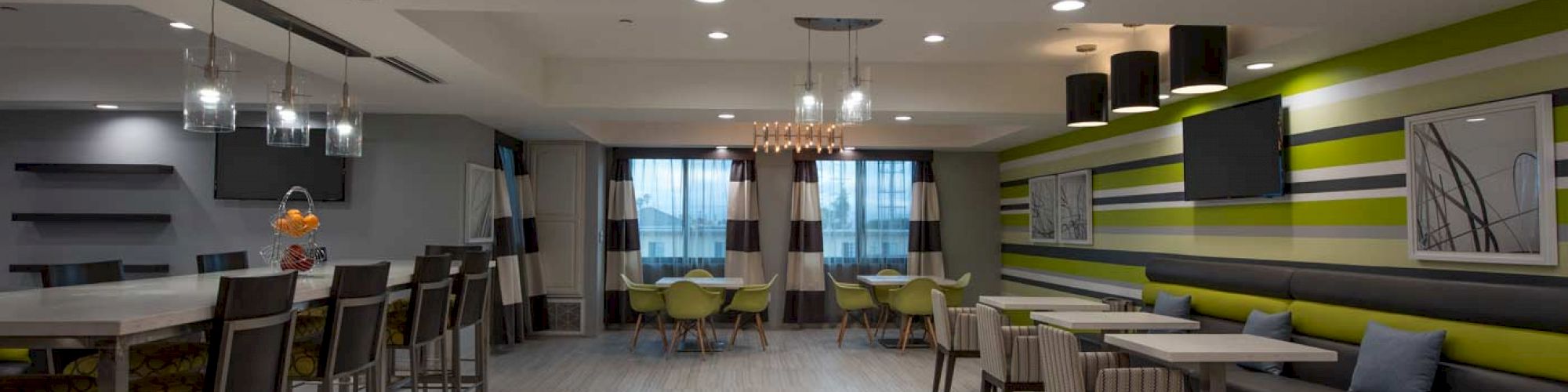 A spacious dining area with modern decor features a row of high chairs at a counter, tables with chairs, a TV, and a contemporary chandelier.