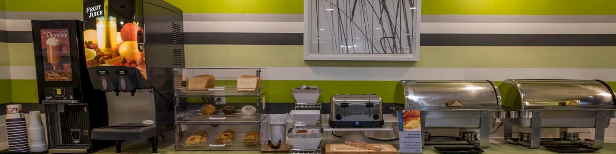 The image shows a breakfast buffet setup with drink dispensers, pastries, and warming trays on a counter under modern lighting.