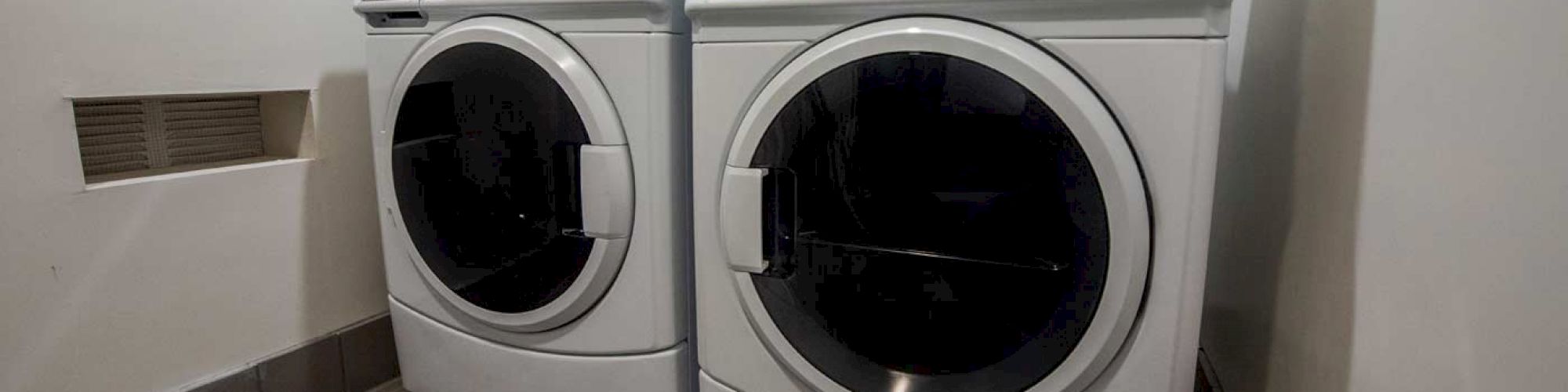 A laundry room with a washer and dryer side by side on a tiled floor against a white wall, with some controls mounted above them.